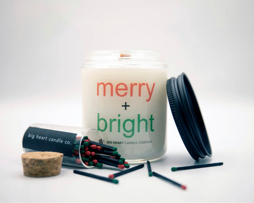 In the center of the photo is clear jar candle with a wood wick and labeled with a merry + bright label in red and green. There is a black metal candle lid and a clear vial with about 35 green and red tipped black stem matches spilling out of it. A small cork lid for the vial is on the far left of the photo.